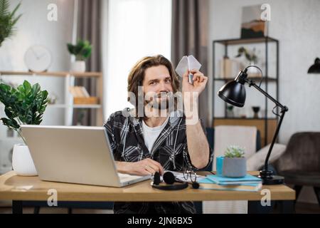Attraktiver kaukasischer Mann in legerem Outfit, der mit einem modernen Laptop am Schreibtisch sitzt und mit einem Papierflugzeug spielt. Mann langweilt sich bei der Arbeit. Unproduktives Konzept. Stockfoto