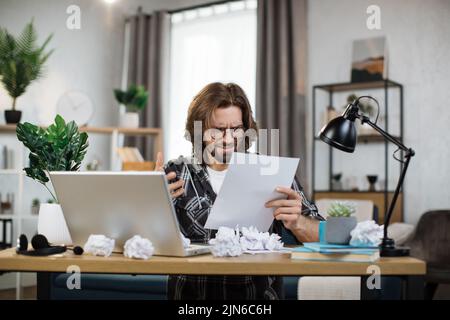 Schockiert junge kaukasische Mann sitzt am Tisch mit einem modernen Laptop im Home Office, lesen Papier Brief Post und Hände zu verbreiten, halten Dokument oder Rechnungen, nachdenklichen Kerl bekam schlechte Nachrichten, Schuldenmitteilung Stockfoto