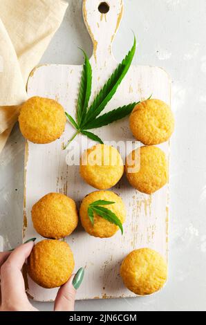 Viele Muffins aus Marihuana mit Cannabisblättern auf einem Holzbrett und eine weibliche Hand nimmt einen Muffin. Glutenfrei. Alternative Süßwaren-Konzept. Hor Stockfoto