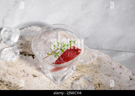Wodka aus Schrotglas mit Eis, dekoriert mit Erdbeerscheibe und Thymian auf Travertin-Podium auf rosa Marmorstein Hintergrund. Alkoholcocktail Shot wi Stockfoto