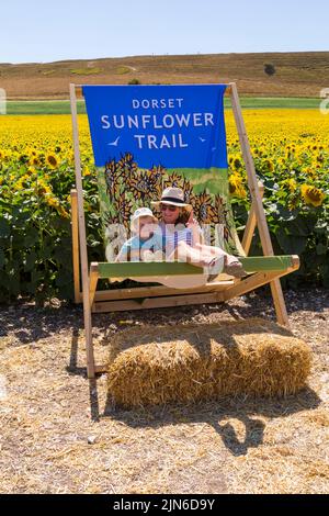 Maiden Castle Farm, Dorchester, Dorset, Großbritannien. 9.. August 2022. Besucher strömen an einem heißen, sonnigen Tag zur Maiden Castle Farm, in der Nähe von Dorchester in Dorset, um den Dorset Sunflower Trail zu erkunden. Die leuchtend gelben Sonnenblumen brauchen glücklicherweise keine regelmäßige Bewässerung, so gedeihen Sie im Sonnenschein und bieten einen fröhlichen Blick gegen die ausgetrockte Umgebung, mit den kontrastierenden gelben Blüten gegen den blauen Himmel, während die Hitzewelle anhält. Amy und Ezra ruhen sich im großen Liegestuhl aus. (Genehmigung vom Bauernhof der Burg Maiden). Quelle: Carolyn Jenkins/Alamy Live News Stockfoto