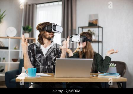 Junge Menschen spielen zu Hause Spiele und meditieren mit VR-Brillen und Laptops. Glückliche Familie genießen künstliche Realität Unterhaltung sich mit Innovationen. Stockfoto