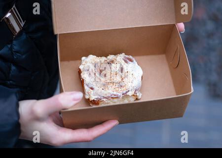 Ein Mädchen hält einen Cinnabon mit Creme in einer Bastelkiste. Lieferung von frischen Backwaren Stockfoto