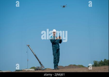 Ein Mann in Helm und Overall steuert eine Drohne auf einer Baustelle. Der Bauherr führt die technische Überwachung durch. Stockfoto