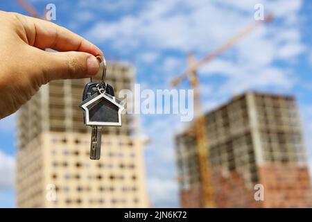 Schlüsselanhänger in Hausform und Schlüssel in männlicher Hand auf dem Hintergrund von Baukräne und Neubauten. Immobilienmakler, Konzept der Kauf einer Wohnung Stockfoto