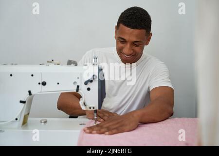 Lächelnder junger Mann, der im Arbeitsatelist Kleidung näht Stockfoto