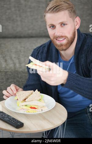 Junger Mann, der zu Hause leckeres Sandwich isst Stockfoto