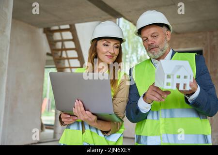 Realtor hält einen offenen Laptop und schaut sich das Modellhaus an Stockfoto