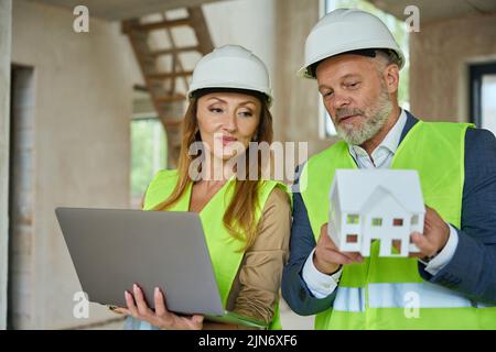 Der Agent hält einen offenen Laptop und sieht sich das Modell des Hauses an Stockfoto