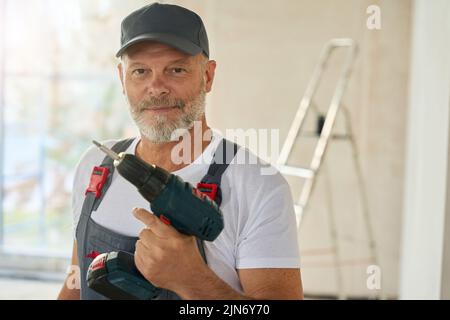 Porträt eines lächelnden bärtigen Arbeiters in Overalls mit Bohrer Stockfoto