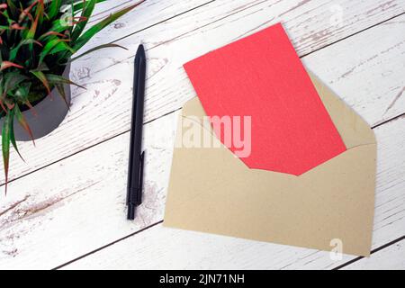Rote Karte Mockup mit Umschlag, Stift und Topfpflanze auf Holztisch. Stockfoto