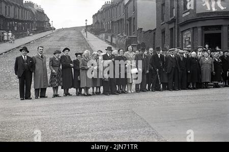 1950s, historisch, Busfahrt, Sheffield-Torquay Express. Gut gekleidete ältere Passagiere stehen auf der Straße, in der Grimesthorpe Road, Pitsmoor, Shefffield, South Yorkshire, England, Großbritannien, für ein Gruppenfoto vor der Reise. Hinter ihnen der steile Hügel der Ditchingham Road. An der Ecke befindet sich das Lebensmittelgeschäft von Lliy Bell. Stockfoto