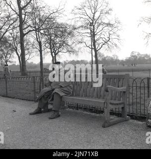 1950s, historisch, ein Gentleman mit Mantel und Hut, der auf einer Holzbank im Londoner Hyde Park, einem großen Royal Park im Zentrum der Stadt, ein Nickerchen macht. Stockfoto