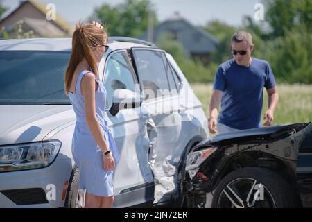 Verärgerte Frauen und Männer, die stark beschädigte Fahrzeuge fahren und argumentieren, wer sich für einen Autounfall auf der Straßenseite schuldig gemacht hat. Straßenverkehrssicherheit und Versicherungskonzept Stockfoto