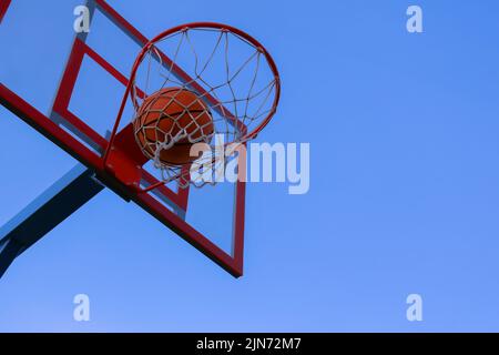 Ein Basketball in einem Netz auf einem blauen Himmel Hintergrund. Der Ball traf den Ring. Basketballplatz im Freien. Der Basketball flog durch den Ring und flog durch den Korb und das Netz hinein. Stockfoto