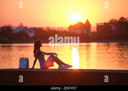 Einsame Frau, die am warmen Abend am Seeufer sitzt. Einsamkeit und Entspannungskonzept