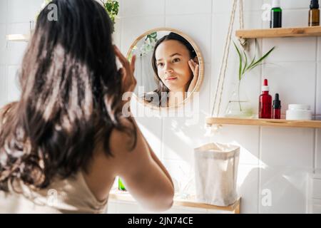 Schöne hispanische Frau, die im Spiegel im weißen, umweltfreundlichen Badezimmer schaut. Holzschnepfe und wiederverwendbare Kosmetikflaschen. Wellness, Hautpflegekonzept Stockfoto