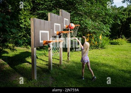 Kleines Mädchen spielt Basketball auf drei Korbständer, wo Reifen auf verschiedenen Höhen sind. Kreative aktive Sportspielgeräte für Kleinkinder Stockfoto