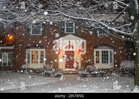 Schneefall auf schönen Backsteinhaus mit Säulen und Erkerfenster mit Weihnachtsbaum Licht und roten Schlitten und Kranz auf der Veranda Stockfoto