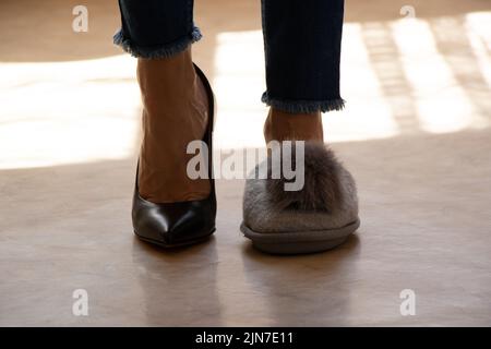 Auf einem Bein mit hohen Absätzen und auf dem anderen Bein hat das Mädchen Hausschuhe mit einem Bubo auf dem Boden des Hauses, verschiedene Schuhe auf den Füßen des Mädchens, Stockfoto