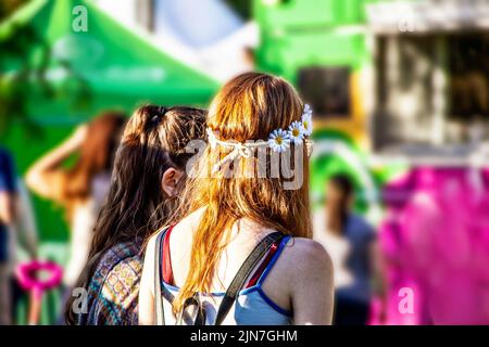 Zwei Mädchen - eines mit roten Haaren und Gänseblümchen-Stirnband - warten an Fasching in der Schlange beim Food Truck - knallige Farben von Pink und Grün Stockfoto