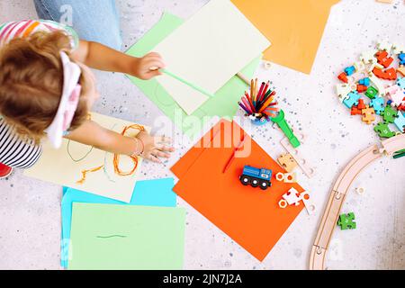 Draufsicht auf ein kleines Mädchen, das auf allen Vieren steht und grünen Bleistift um Buntstifte, Spielzeug und Puzzles im Klassenzimmer hält. Stockfoto