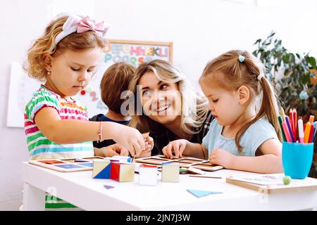 Porträt einer jungen glücklichen Frau, die mit Buntstiften Brettspiele am Tisch sitzt. Lehrer, der ein ernsthaftes kleines Mädchen ansieht. Stockfoto