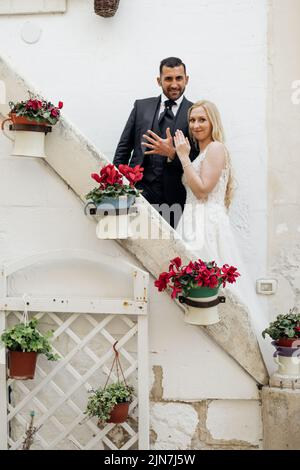 Porträt eines Ehepaares, das auf einer Treppe mit hängenden Blumentöpfen steht, die Kamera anschaut und die Hände mit Ringen zeigt. Stockfoto