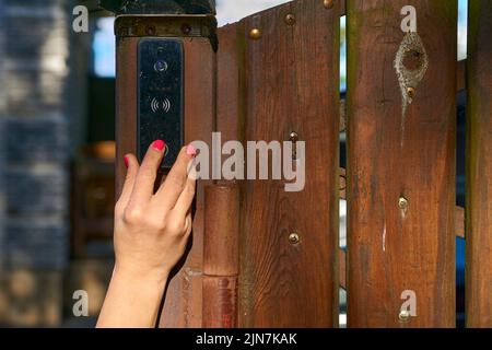 Die Hand einer Frau drückt eine Türklingel auf einen Holzzaun im Garten Stockfoto