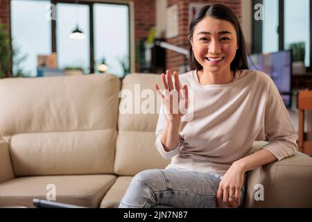Glückliche junge asiatische Chineese filipino Frau sitzt auf dem Sofa zu Hause halten Telefon am Bildschirm winkende Hand Video Anruf Entfernung Freund online in mobilen Chat-App mit Smartphone-Videochat-Anwendung Stockfoto
