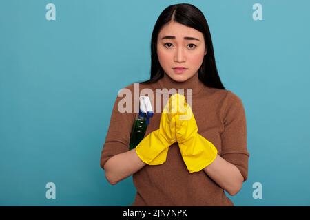 Junge verängstigte asiatische Hausfrau legte ihre Hände zusammen mit gelben Handschuhen und einem Waschmittelsprüher unter dem Arm isoliert vor einem blauen Hintergrund, Hausfrau Arbeiter, Reinigung Haus Konzept, Stockfoto