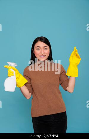 Zufrieden lächelnd asiatische Hausmädchen halten Spray gelben Gummihandschuh isoliert auf einem blauen Hintergrund, Reinigung Home Konzept, lächelnd Gesichtsausdruck weiblich bereit, Hausaufgaben zu tun Stockfoto