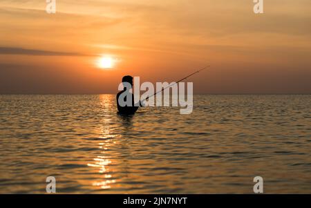 Ein eingeflügelter Fischer ist im orangefarbenen Glanz eines tropischen Sonnenuntergangs silhouettiert, als er im Wasser steht und seine Angelköder abwirft Stockfoto