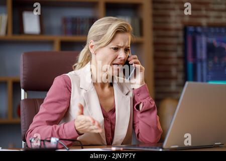 Seriöse Geschäftsfrau, die unangenehme Telefongespräche mit einem Laptop im Büro geführt hat. Weibliche Trader hören schlechte Nachrichten, fühlt sich verzweifelt Stockfoto
