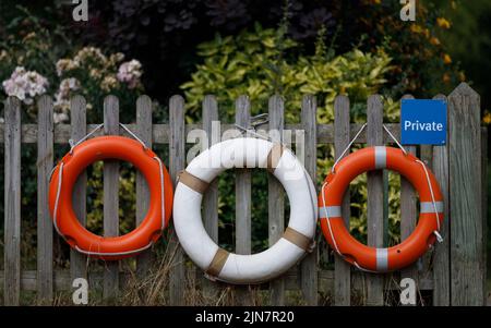 Drei Rettungsbojen hängen an einem Holzzaun mit Blumen und Sträuchern dahinter. Orange und weiß, privat Stockfoto