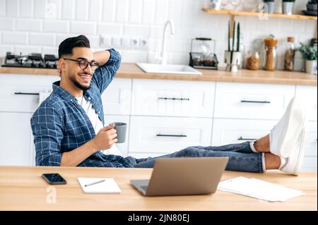 Entspannter indischer oder arabischer moderner Mann in stilvoller Kleidung, sitzen zu Hause in einer Küche mit den Beinen auf dem Tisch, halten eine Tasse Kaffee in der Hand, arbeiten fernab von zu Hause mit Laptop, chillen, lächeln Stockfoto