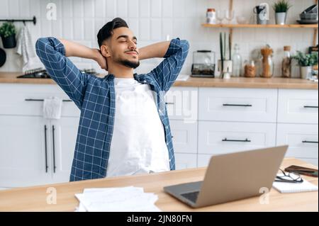 Entspannter, ruhiger araber oder inder, Freiberufler, arbeitet von zu Hause aus, sitzt in der Küche am Arbeitsplatz, macht eine Pause von der Arbeit, legt seine Hände hinter den Kopf, schließt seine Augen, träumt von Ruhe, lächelt Stockfoto