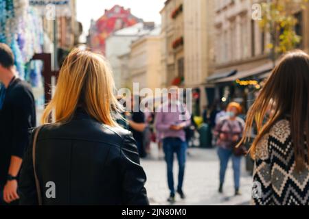 Unschärfe zwei stilvolle blonde und weiße Frauen, die in schwarzer Jacke in der herbstlichen Stadtstraße bei Sonnenuntergang im Freien spazieren. Blick von hinten. Freundschaft Stockfoto