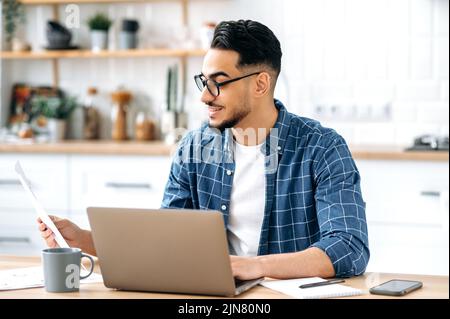 Job mit Dokumenten. Intelligenter erfolgreicher arabischer oder indischer Mann mit Brille, Freiberufler oder Büroangestellter, der aus der Ferne arbeitet, sitzt an einem Schreibtisch in der Küche, arbeitet an einem Projekt, analysiert Dokumente, lächelt Stockfoto