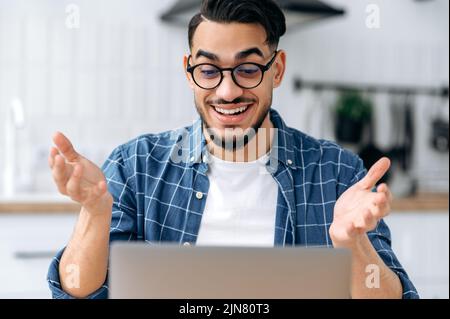 Aufgeregter, positiver arabischer oder indischer Mann mit Brille, cleverer Student oder Freiberufler, Fernkommunikation per Videoanruf per Laptop, Online-Webinar anhören, Vortrag, lächelnd freundlich Stockfoto