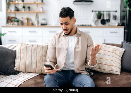 Ein verwirrter inder oder araber in legerer Kleidung, sitzt auf einem Sofa in einem Wohnzimmer, hält ein Smartphone in der Hand, schaut verwirrt auf das Handy, breitet seine Arme aus, liest unerwartete Nachrichten Stockfoto