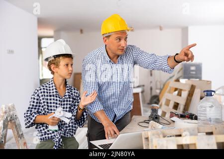 Ein Architekt und ein Junge verwenden einen Laptop, während sie über den Arbeitsprozess im Haus sprechen, der gerade renoviert wird Stockfoto