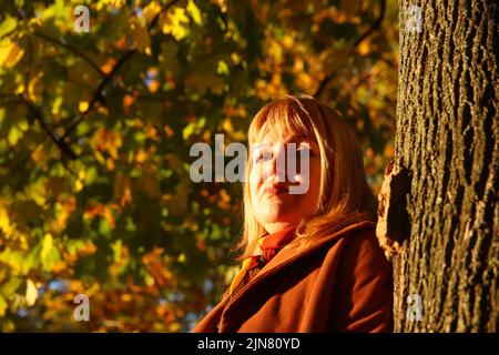 Unschärfe-Porträt der hübschen kaukasischen blonden Frau in bunten gelben Ahorn Herbst Park. Helle stilvolle Frau im Mantel in der Nähe Baumstamm. Hallo Herbstkonz Stockfoto