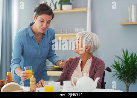 Orangensaft ist das Beste für Sie. Eine glückliche ältere Frau, die zu Hause sitzt und mit ihrer attraktiven jungen Tochter frühstückt. Stockfoto