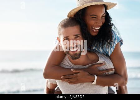 Liebe ist so ein glückseliges Gefühl. Porträt eines jungen Mannes, der seine Freundin am Strand mit Huckepack zurückkickt. Stockfoto