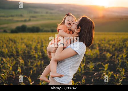 Porträt einer kaukasischen jungen Mutter und ihrer Tochter auf einem Maisfeld, das sich umarmt und küsst. Glückliche Familie, Kindheit. Mutter Natur. Frohe Familie Stockfoto