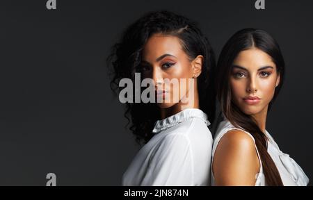 Alles an ihnen ist atemberaubend. Porträt zweier attraktiver junger Frauen, die sich vor dunklem Hintergrund in einem Studio Rücken an Rücken posieren. Stockfoto
