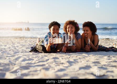 Selfie-Zeit. Ein attraktives junges Trio von Frauen, die sich am Strand niederlegen und tagsüber Selfies zusammen machen. Stockfoto