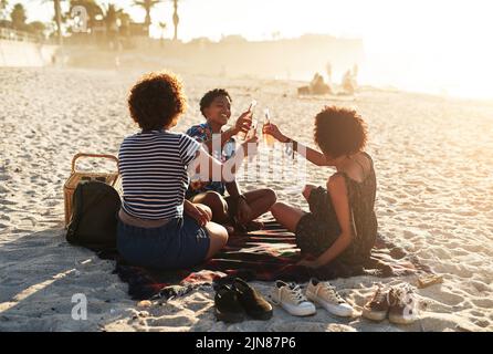 Sonne, Getränke, Freunde und der Strand sind alles, was wir brauchen. Ein attraktives junges Trio von Frauen feiern und einen Drink zusammen am Strand während der Stockfoto