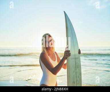 Das Leben ist einfach besser, wenn man surft. Ein Porträt einer attraktiven jungen Frau, die in einem Badeanzug mit einem Surfbrett am Strand steht. Stockfoto